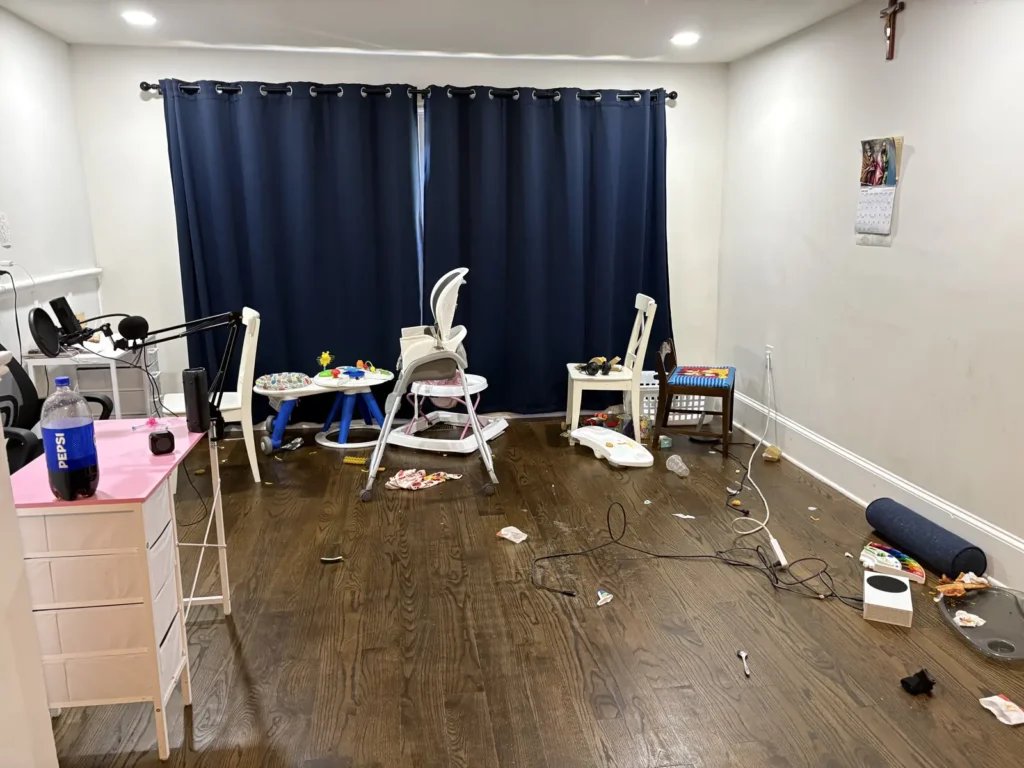 Cluttered playroom before organizing, with baby gear, loose cords, and food wrappers scattered across the hardwood floor