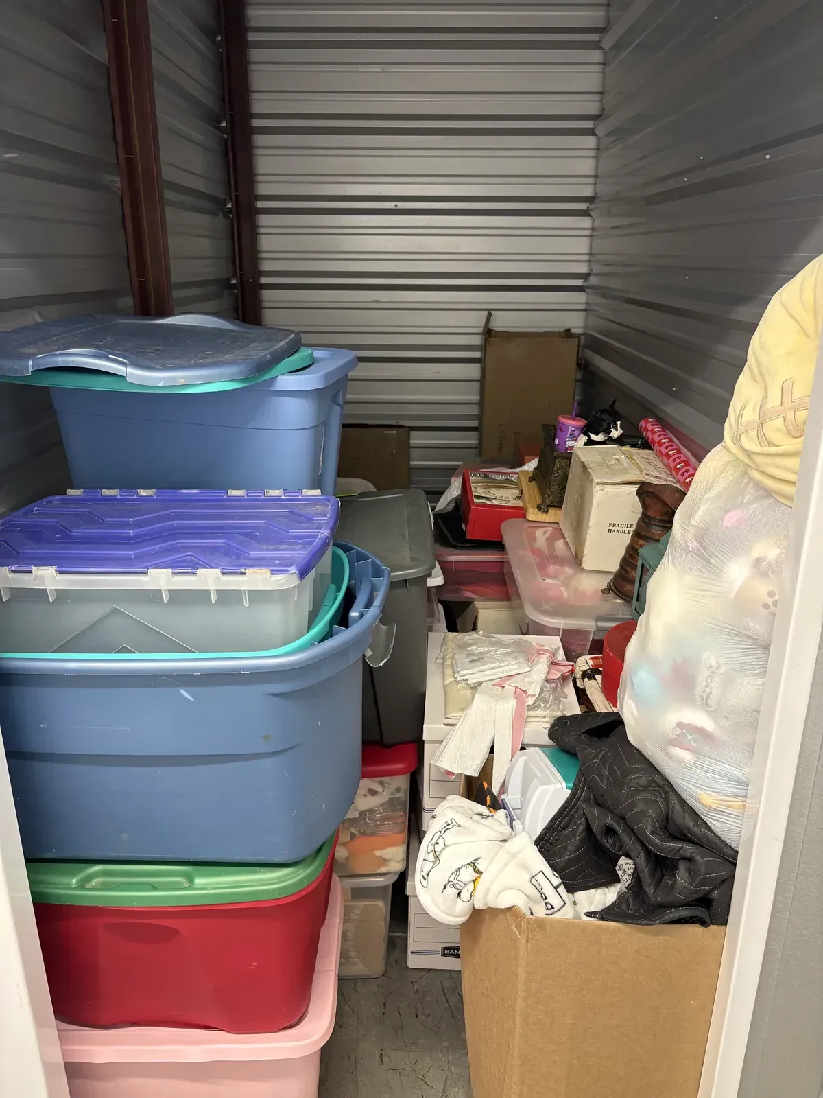 Cluttered self-storage unit before organizing, with mismatched bins stacked to the ceiling and a garment bag wedged against the wall